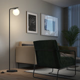 A modern living room with a green armchair, beige sofa, floor lamp with a round white shade, framed wall art, a potted plant, and a rug on wooden flooring. The lamp is turned on, creating a warm light.