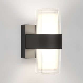 The Artika Colton modern LED indoor-outdoor wall light 5 CCT features a cylindrical frosted glass shade within clear glass, secured by a matte black metal band and rectangular base, and is shown mounted on a light-colored wall.