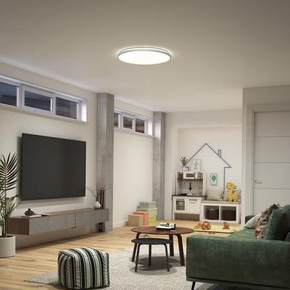 A modern living room with a green sofa, round coffee table, large wall-mounted TV, pouf, wall plants, play kitchen set, and an Artika Jupiter 23 in. LED flush mount light with remote installed on the ceiling above wooden floors and high windows.