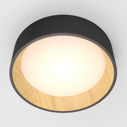 The Artika Alton LED flush mount features a round black outer frame, wooden inner rim, and glowing white center—modern lighting for any room. Seen here from below against a plain white ceiling.