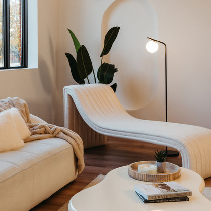 A modern living room features a sculptural cream chaise lounge, a round white coffee table with books and tray, a cozy sofa with cushions and blanket, the Artika Clayton dimmable LED modern mid-century floor lamp, and large green plants by a curved wall niche.
