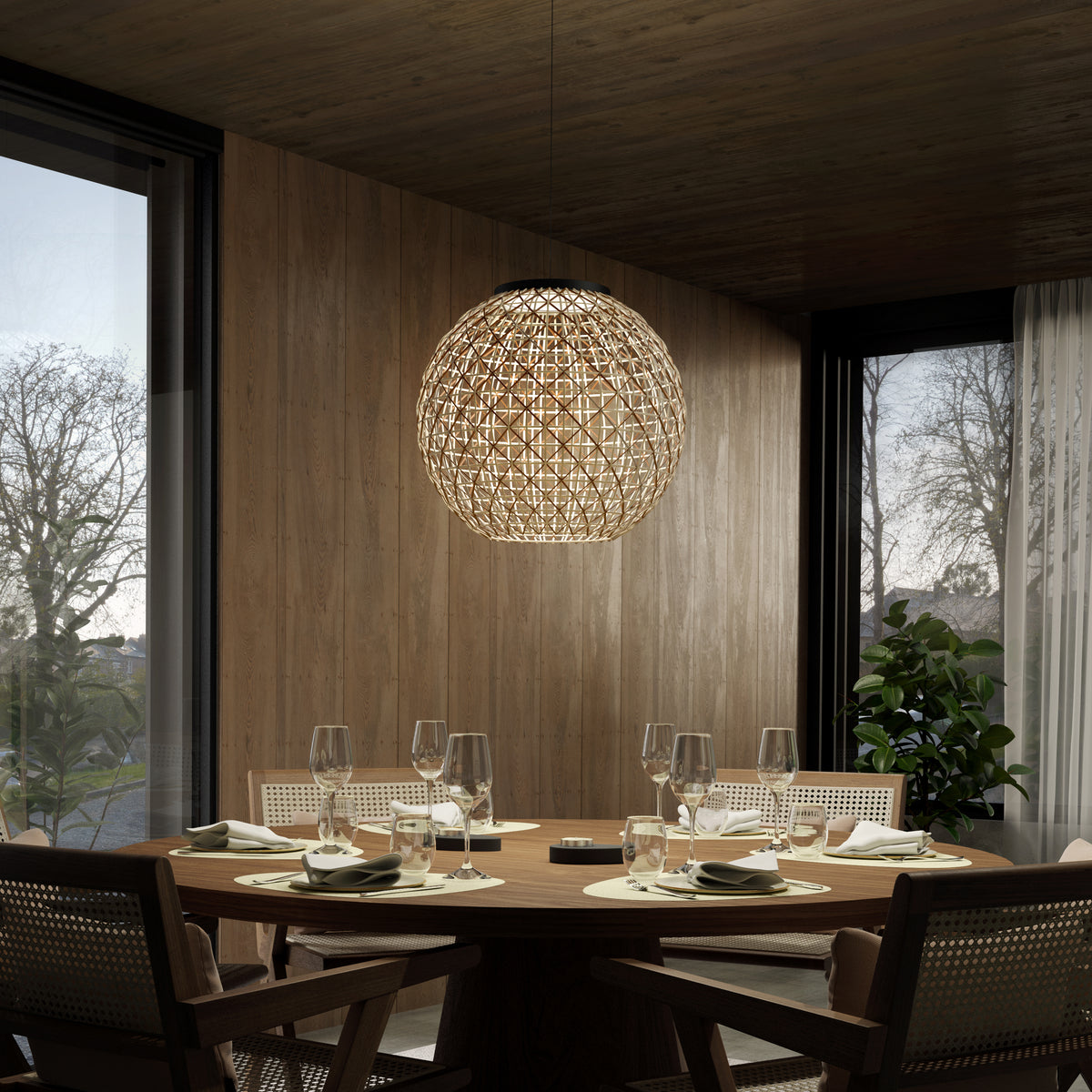 A round wooden dining table set for six with glassware and plates sits beneath a large, woven pendant light in a modern, wood-paneled dining room with large windows and natural light.