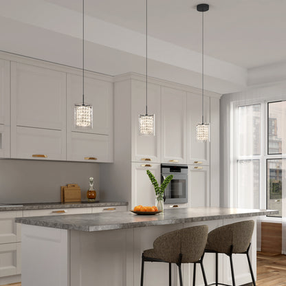 Modern kitchen with white cabinets, a large island featuring a gray marble countertop, two cushioned bar stools, three Artika Crystal Cube 1-light LED pendant lights, a vase with green leaves, and a bowl of oranges on the counter.