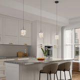 Modern kitchen with white cabinets, gray marble countertop island, two upholstered barstools, three pendant lights, a vase with green leaves, and a bowl of oranges. Sunlight streams in through large windows.