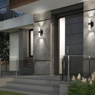 Modern house entrance with gray walls, a sleek dark front door, glass railing, concrete steps, and Artika Gemini indoor-outdoor LED wall lights on either side of the door, plus small plants by the steps.