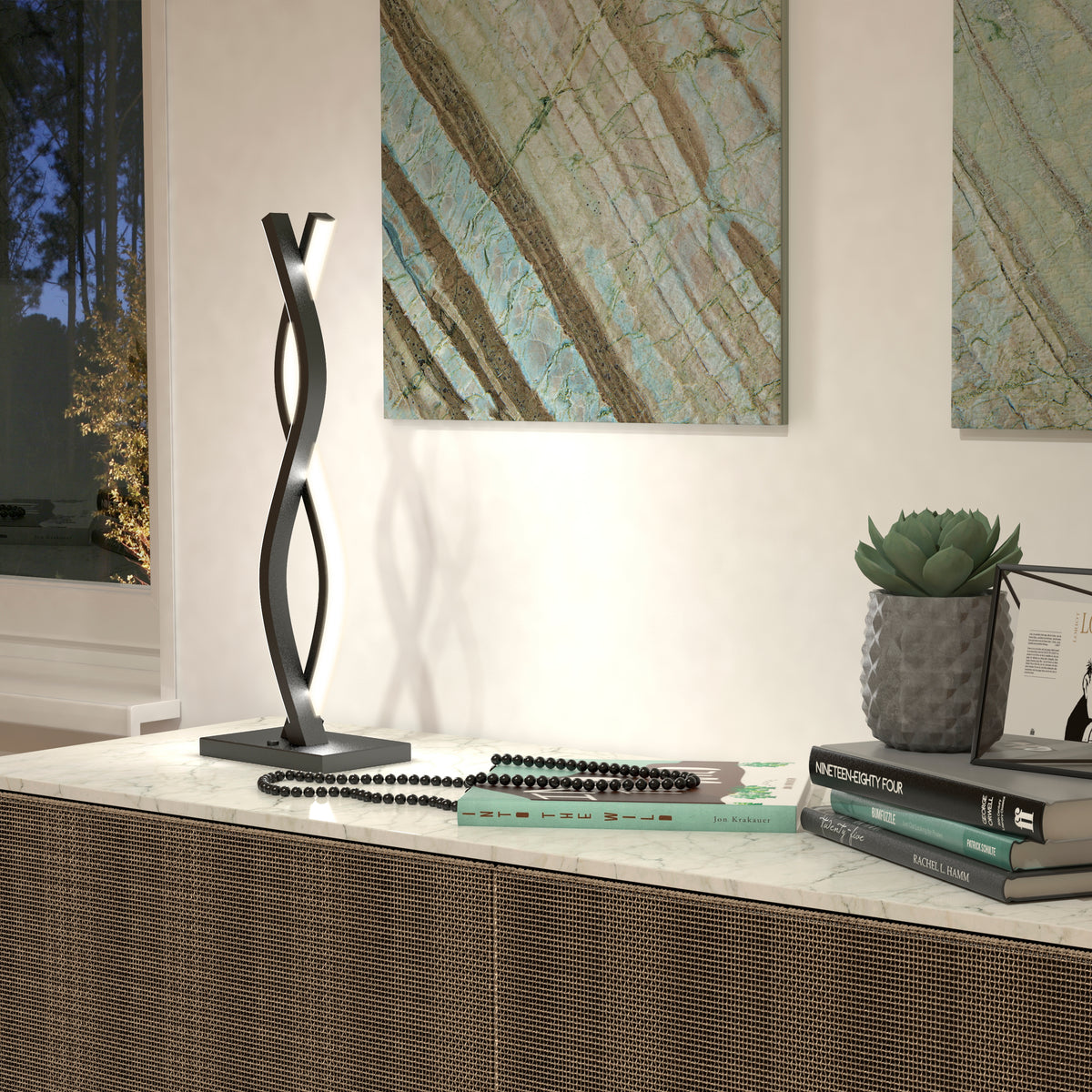 A modern table lamp with a twisted metal design sits on a marble surface, alongside books, a black beaded necklace, a small potted succulent, and abstract art hanging on the wall in the background.