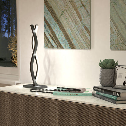 A modern table features the Artika Wave LED Table Lamp with twisted black metal, a stack of books, a beaded necklace, a succulent in a textured pot, and two abstract art pieces on a neutral wall in the background.