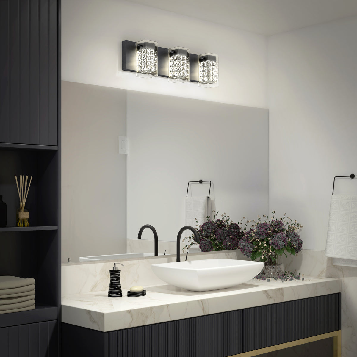 A modern bathroom with a square white sink on a marble counter, a black faucet, decorative soap, a bouquet of purple flowers, folded towels, and a three-light vanity fixture above a large mirror.