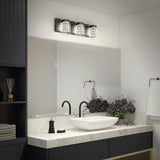A modern bathroom with a square white sink on a marble counter, a black faucet, decorative soap, a bouquet of purple flowers, folded towels, and a three-light vanity fixture above a large mirror.