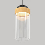 A modern pendant light with a cylindrical clear glass shade, a wooden accent at the top, and a black metal fixture, suspended against a plain gray background.