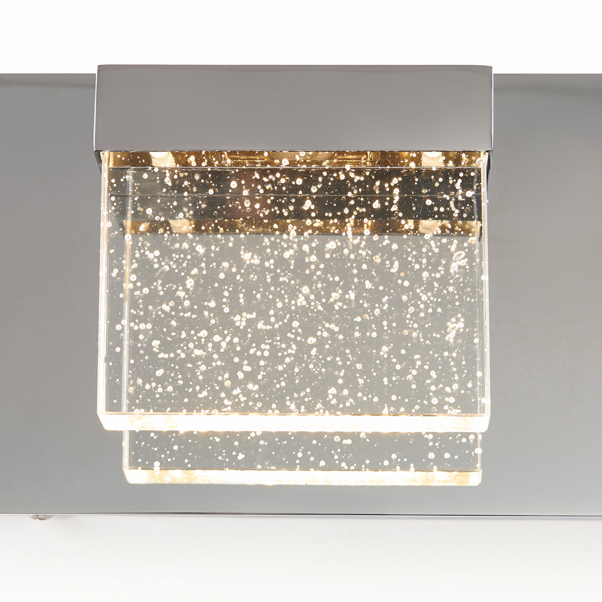 A modern square wall light fixture with a metallic base and a clear acrylic shade embedded with bubbles, illuminated and glowing warmly.