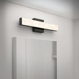 A modern rectangular wall sconce with a textured, illuminated front panel is mounted above a mirror on a white wall in a contemporary bathroom.