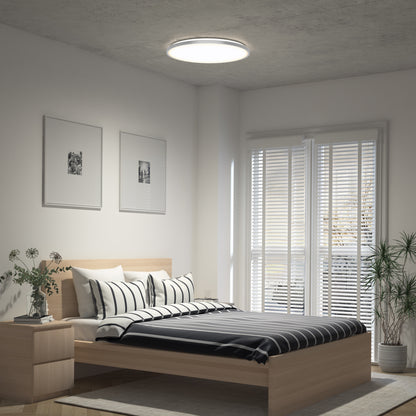A modern bedroom features a wooden bed with black and white striped bedding, two framed wall pictures, a nightstand with flowers, large windows with blinds, potted plants, and an Artika Jupiter 23 in. LED flush mount light with remote on the ceiling.