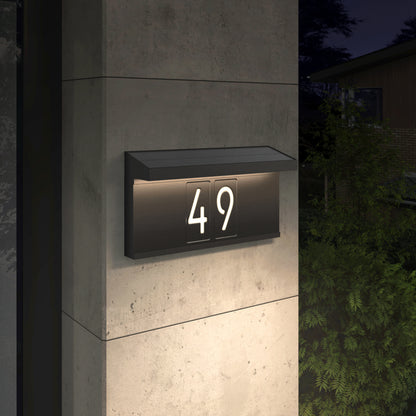 The Artika Bowen solar LED outdoor address wall light 5 CCT displays the number 49 on a concrete wall at night, offering maintenance-free illumination, with greenery and another building visible in the background.
