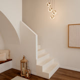 A minimalist interior with white stairs, modern pendant lights, a lantern with candles, a wooden bench with cushions, and a textured wall art piece on a neutral-colored wall.