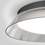 Close-up view of a modern ceiling light fixture with a black base and a transparent, circular acrylic or glass shade, emitting a soft, diffused white light against a neutral background.