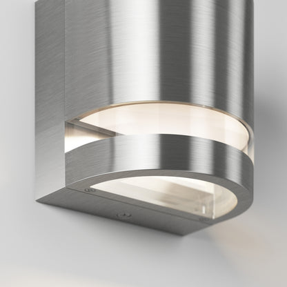 The Artika Valor Round LED indoor-outdoor wall light features a modern, curved brushed metal design with frosted glass, mounted on a light gray wall and emitting light from both the top and bottom.