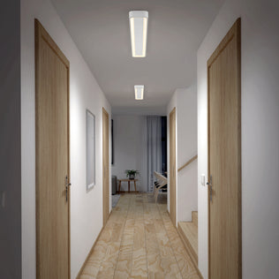 A well-lit hallway with light wooden floors and four closed wooden doors, two per side. Artika's Aiden 48 in. LED modern flush mount ceiling light brightens the space. At the end, a room features a plant, chair, and large curtain.