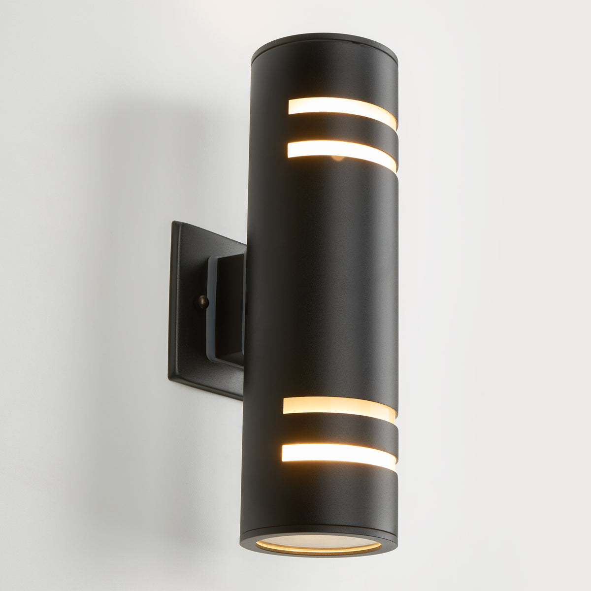 The Artika V3 indoor-outdoor wall light is a modern black cylindrical sconce with horizontal cutouts that emit light from the top, bottom, and cutouts. It’s mounted on a rectangular base against a white wall.