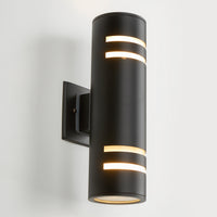 The Artika V3 indoor-outdoor wall light is a modern black cylindrical sconce with horizontal cutouts that emit light from the top, bottom, and cutouts. It’s mounted on a rectangular base against a white wall.