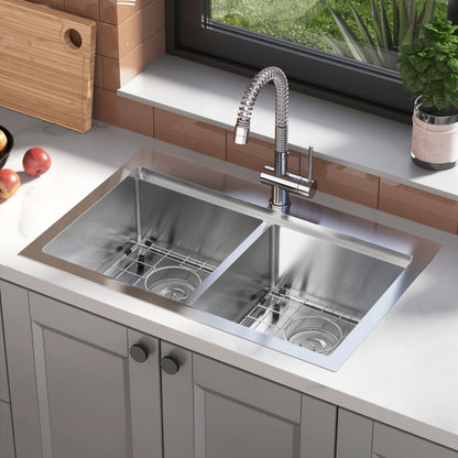 The Artika Newport double bowl stainless steel sink features two basins, a tall flexible faucet, and a white countertop. Nearby are apples, a cutting board, and a potted plant, with a window above the sink.