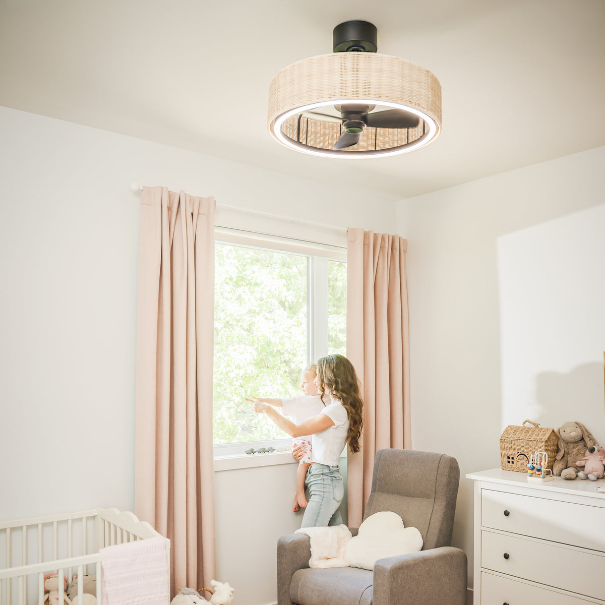 A woman with a baby stands by a sunlit nursery window beneath the Artika Kalani 24 in. LED modern fandelier ceiling fan with light.
