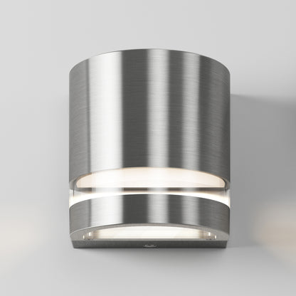 The Artika Valor Round LED indoor-outdoor wall light features a modern cylindrical design in brushed stainless steel with frosted glass, casting up-and-down light on a plain, light gray wall.