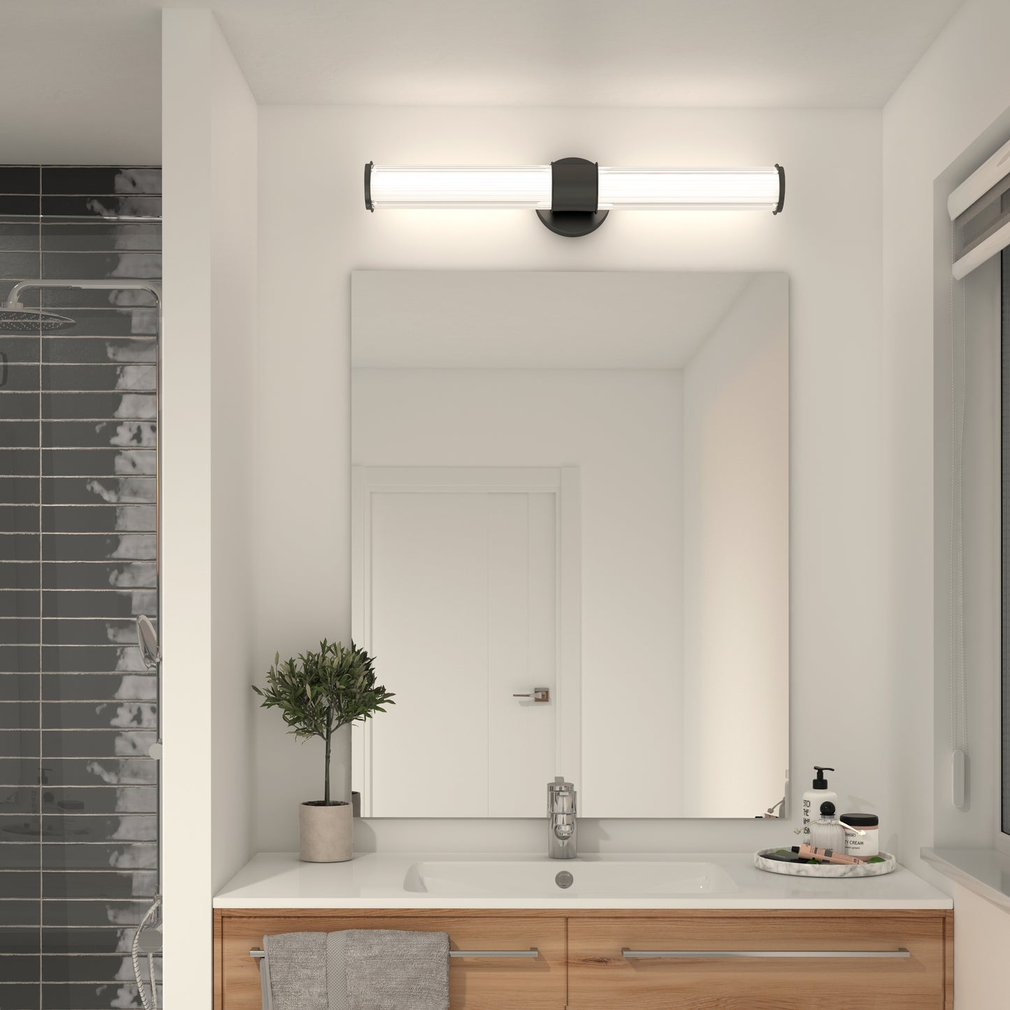 A modern bathroom features a large mirror, wooden vanity, rectangular sink, potted plant, toiletries, and the Artika Kendra LED contemporary bathroom vanity light 3 CCT above the mirror. A gray tiled shower is partially visible on the left.