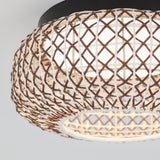 Close-up of a ceiling light fixture with a woven, lattice-style shade made of brown and white strips, allowing light to diffuse through the geometric pattern. The background is plain and light-colored.