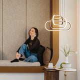 A woman sitting on a cushioned bench, smiling while talking on the phone. She is barefoot, wearing jeans and a black top. Modern hanging lights and a small table with a plant and cups are nearby.