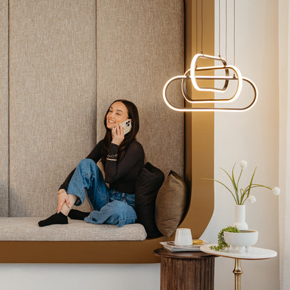 A woman in a black top and jeans smiles while talking on her phone, sitting on a cushioned bench. Above her hangs the Artika Lewis modern LED pendant light 3 CCT. A small table with decor is nearby.