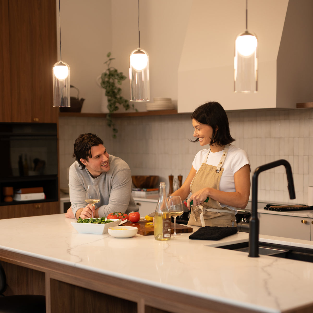 A woman prepares food at a kitchen island while a man leans on the counter, smiling at her. Various vegetables, a salad bowl, and a wine bottle are on the counter under warm pendant lights.