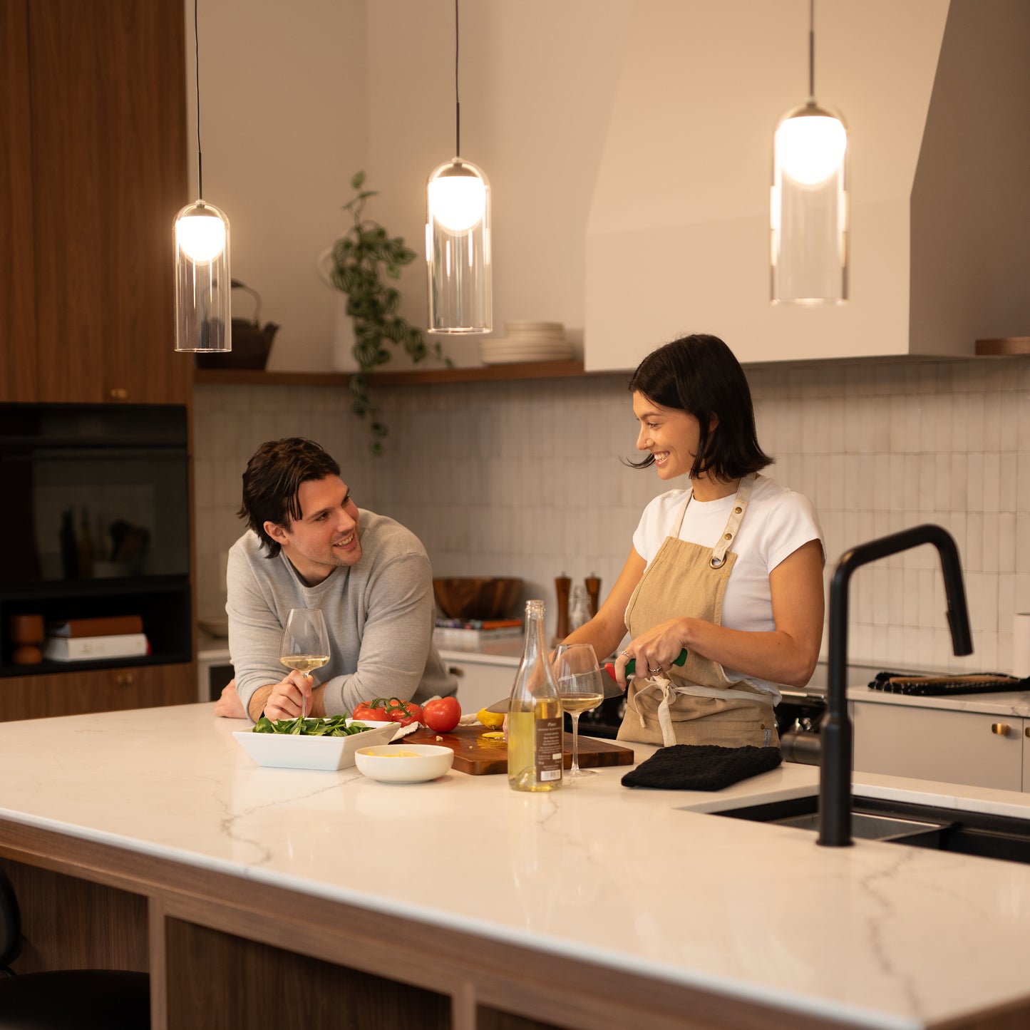Under the Mirah LED modern mid-century pendant light by Artika, a woman in an apron smiles as she chops vegetables at a kitchen island; fresh ingredients and white wine sit nearby in the bright, stylish kitchen.