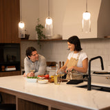 A woman prepares food at a kitchen island while a man leans on the counter, smiling at her. Various vegetables, a salad bowl, and a wine bottle are on the counter under warm pendant lights.