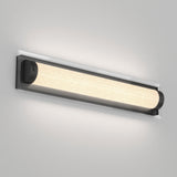 A modern wall-mounted light fixture with a long, cylindrical frosted glass shade and black end caps, emitting a soft, warm glow against a light gray wall.