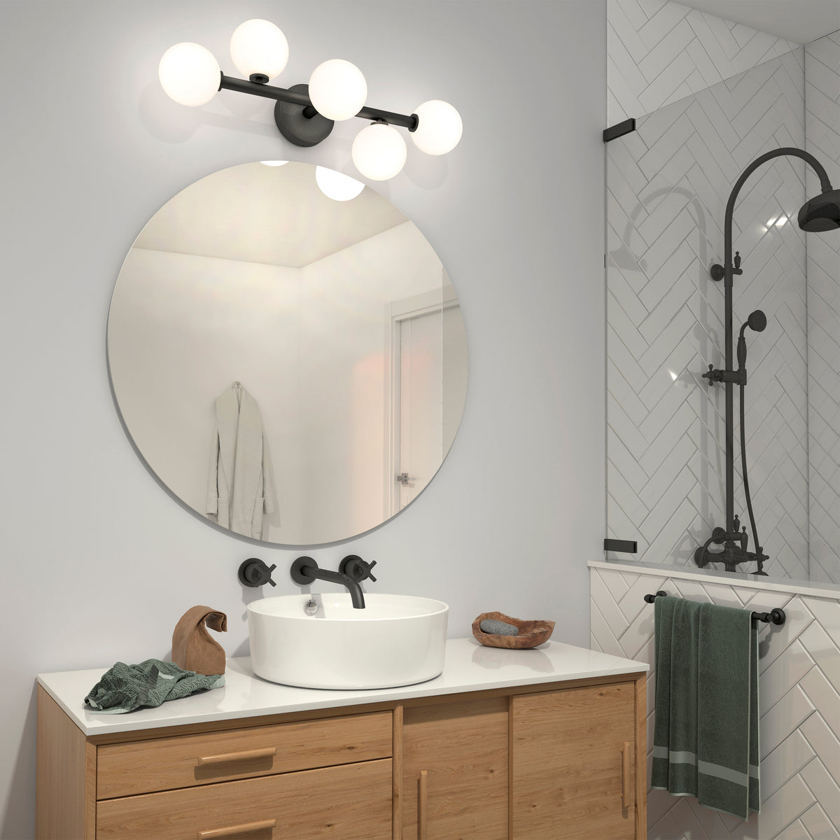 A modern bathroom with a round mirror above a wooden vanity, a vessel sink, wall-mounted faucet, globe light fixture, and a glass shower with black fixtures. A green towel hangs by the shower.