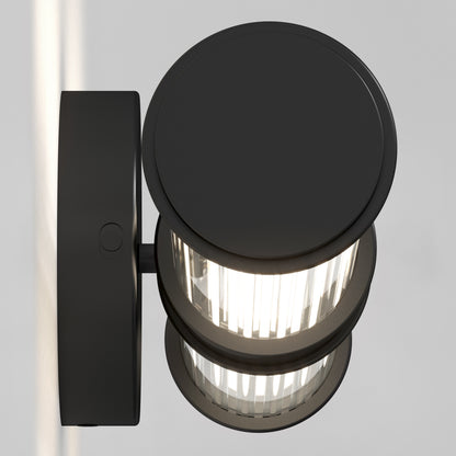The Artika Kendra LED contemporary bathroom vanity light 3 CCT features a modern round black design and vertical ribbed glass shade, mounted with a short horizontal arm, casting light against a plain light gray wall.