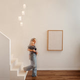 A woman wearing jeans stands at the base of a white staircase holding a young child. Modern pendant lights hang above, and abstract art is mounted on the white wall beside them. The floor is wooden.