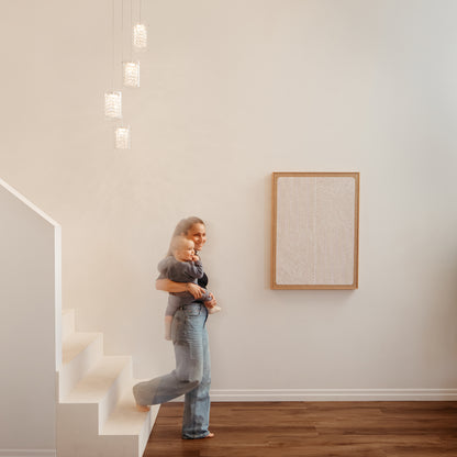 A woman holding a young child stands barefoot at the bottom of white stairs in a minimal, modern room with wooden floors and a Crystal Cube 4-light LED pendant light by Artika overhead.