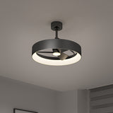 A modern black ceiling fan with an integrated circular LED light is mounted on a white ceiling in a minimalist room with gray walls and a window with horizontal blinds.