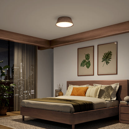 A modern bedroom features a wooden bed, earthy-toned bedding, two plant-themed wall pictures, large city-view window, indoor plants, and is illuminated by the Artika Dunham 15 in. LED flush mount light with 5 CCT settings.