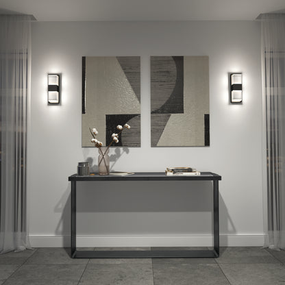 A modern entryway features a black console table, minimalist decor with a vase of cotton stems, stacked books, abstract wall art, and is flanked by Artika's Mazza Frosted indoor-outdoor LED wall lights for sleek contemporary lighting.