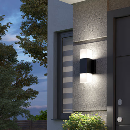 The Artika Lennox indoor-outdoor LED wall light 3 CCT in matte black is mounted by a window with striped blinds, casting adjustable white light on a concrete wall near the front door at dusk. This modern fixture suits versatile lighting needs.