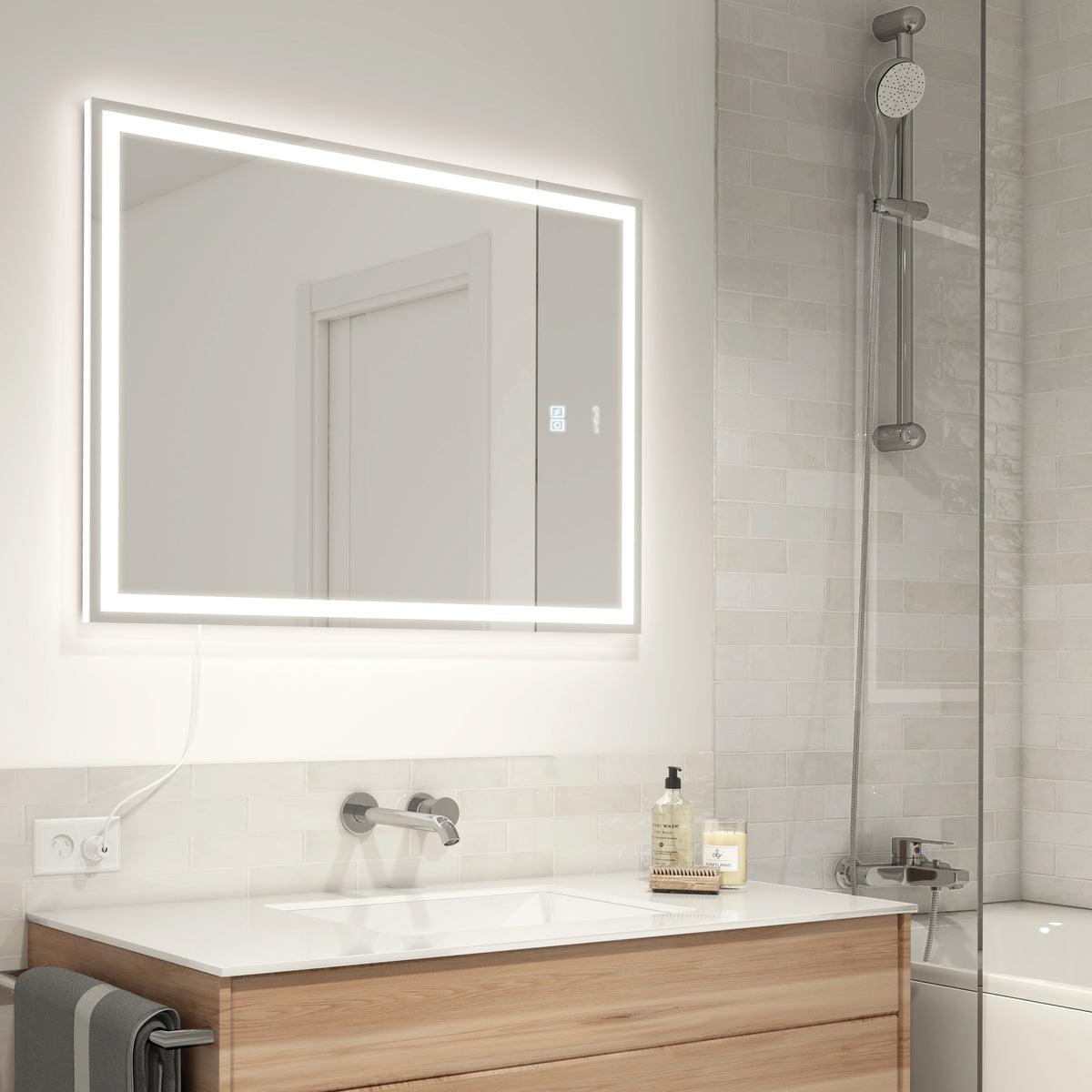 Modern bathroom with a large, illuminated rectangular mirror above a white sink and wooden vanity. A glass shower with a chrome showerhead is on the right. The walls are tiled in light gray.