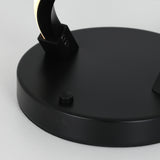 A close-up of a black circular lamp base with a small round button and a curved arm attached, set on a light gray surface.