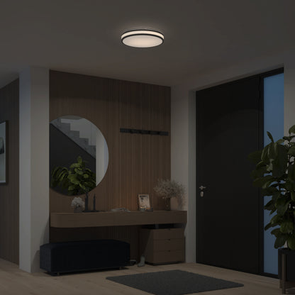 A minimalist entryway is softly lit by the Artika Hemisphere 15 in. LED flush mount light with 5 CCT, and features a round mirror above a wooden console table, decorative pieces, a plant, and a dark front door.
