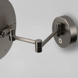 A close-up of a modern, metallic wall-mounted adjustable mirror arm with round joints and a circular wall plate, set against a plain light gray background.