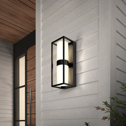 The Artika Sherman modern LED indoor-outdoor wall light 5 CCT features a black metal frame and frosted glass, shown mounted on light gray horizontal siding near a wooden door with green foliage nearby.