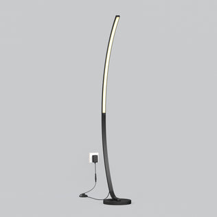 The Artika Arc LED Floor Lamp features a slim, curved black design with integrated LED lighting, standing plugged into a wall outlet against a plain light gray background.