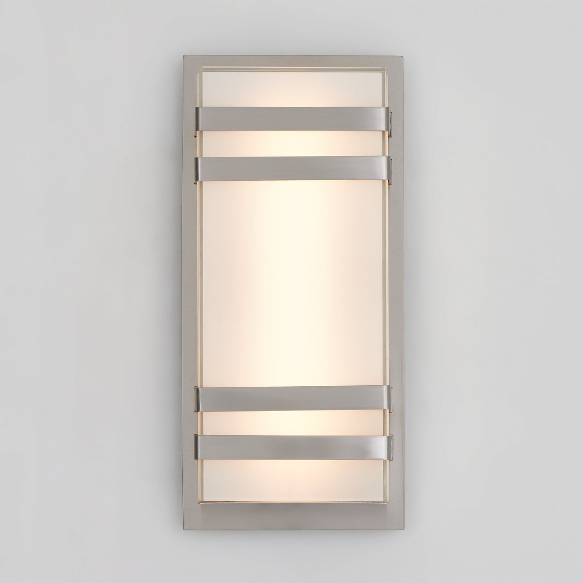 A modern rectangular wall sconce with a frosted glass panel, framed by a metallic border and three horizontal metal bars across the front, emitting soft, diffused light against a plain light gray wall.